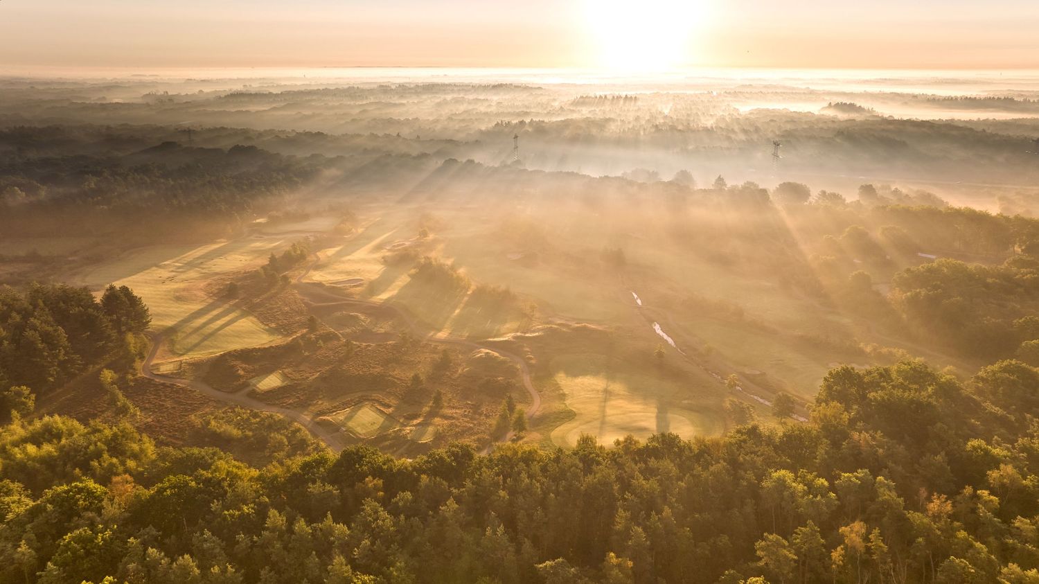 Golden sunrise rays pierce through morning mist over a vast golf course landscape with rolling hills.