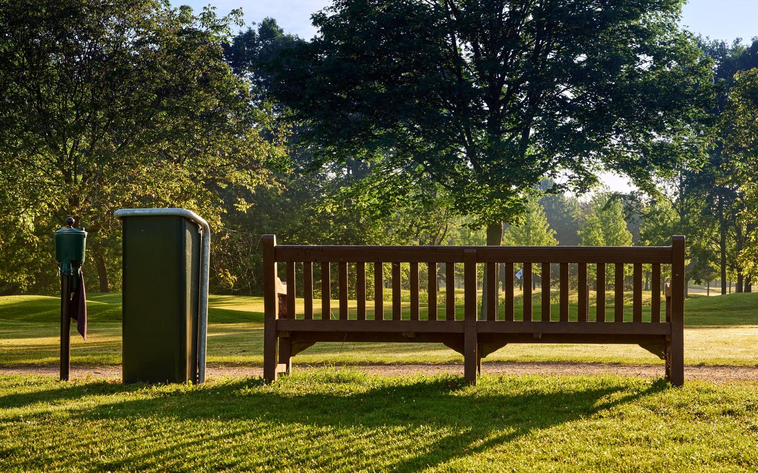 Wooden park bench and waste bin on a sunny golf course with large shade trees in background.
