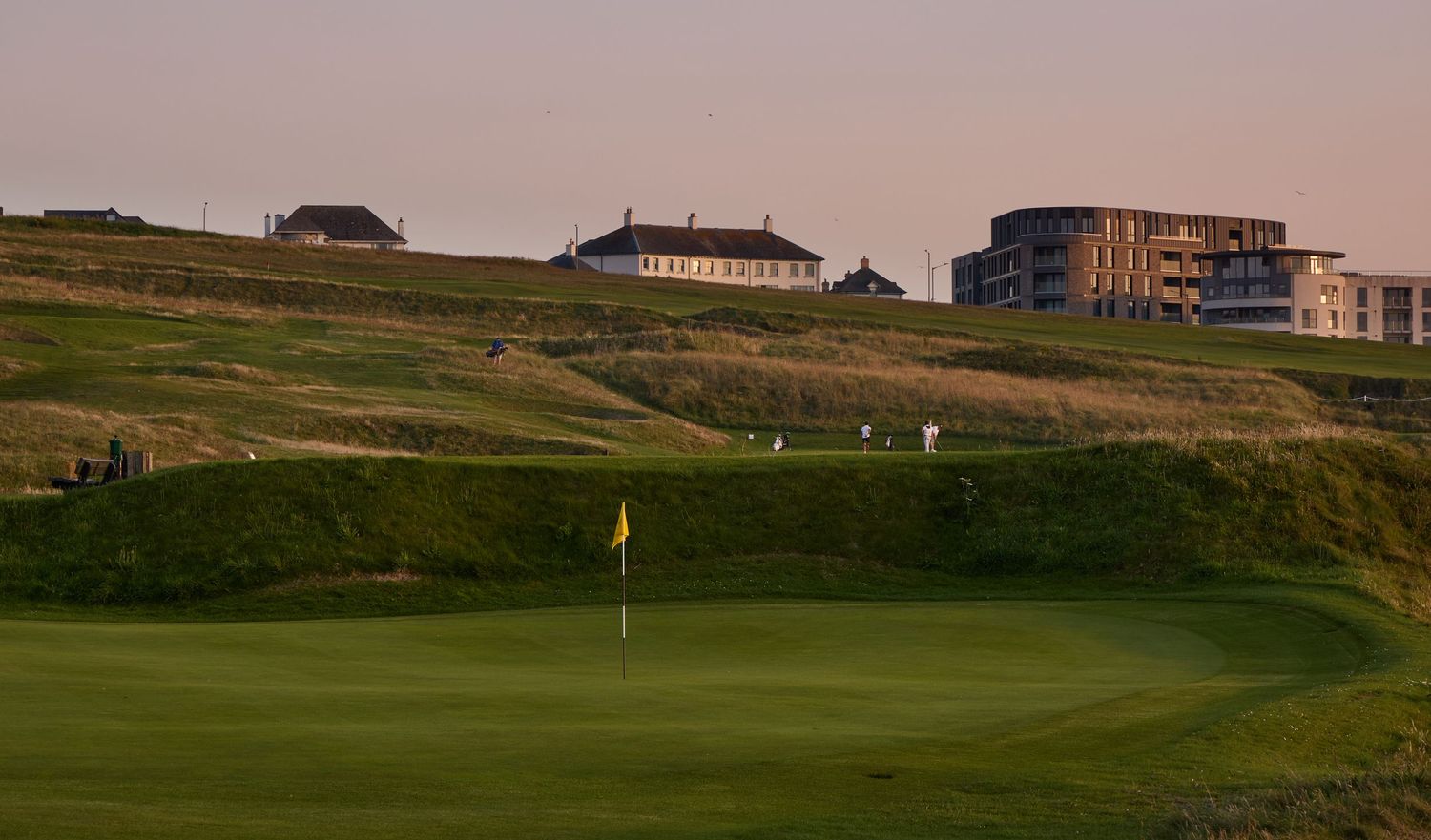 Golf course green with yellow flag near a links-style clubhouse and modern hotel buildings at dusk.