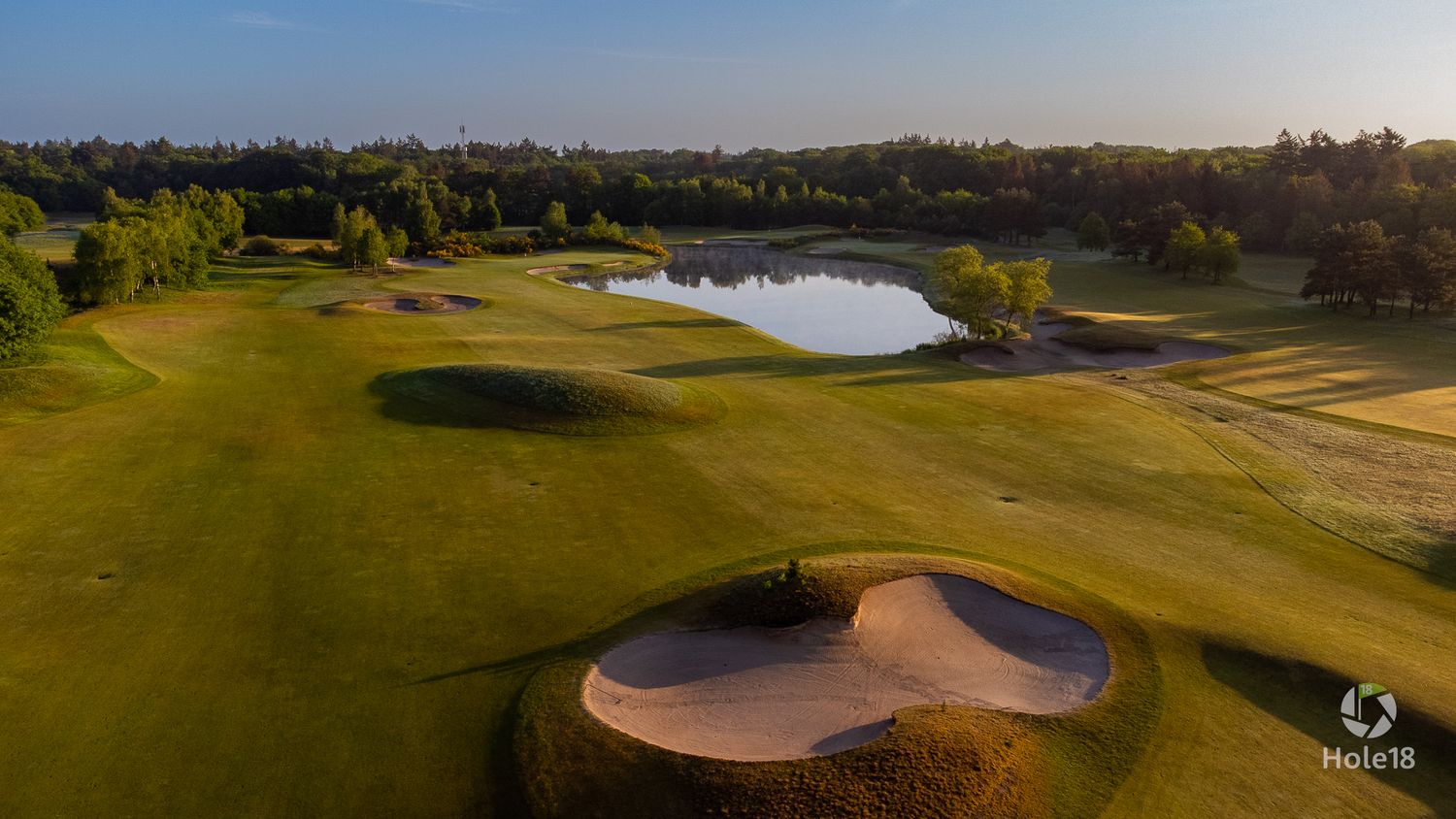 Aerial view of a golf course bunker near a pond reflecting golden sunset light among fairways.