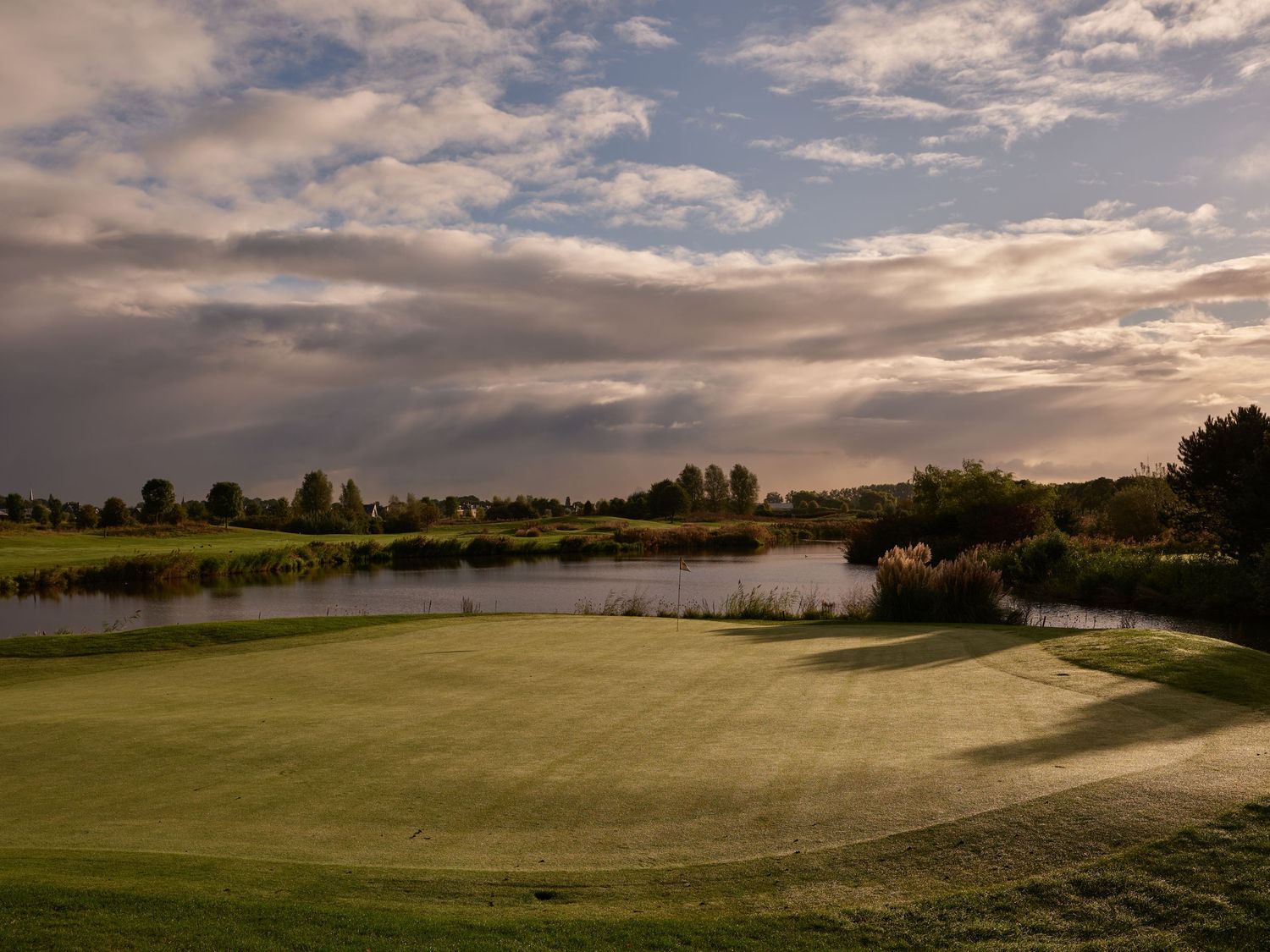 Scenic golf course green beside a calm pond with dramatic storm clouds forming over the landscape at dusk.