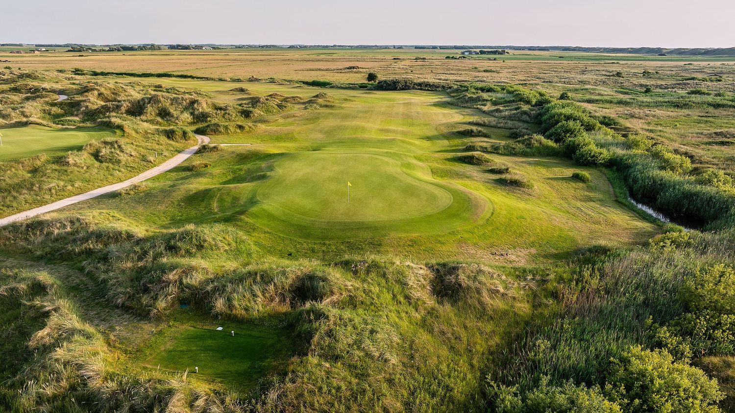 Aerial view of a links golf course with undulating fairways, natural dunes, and wild grass under a clear sky.