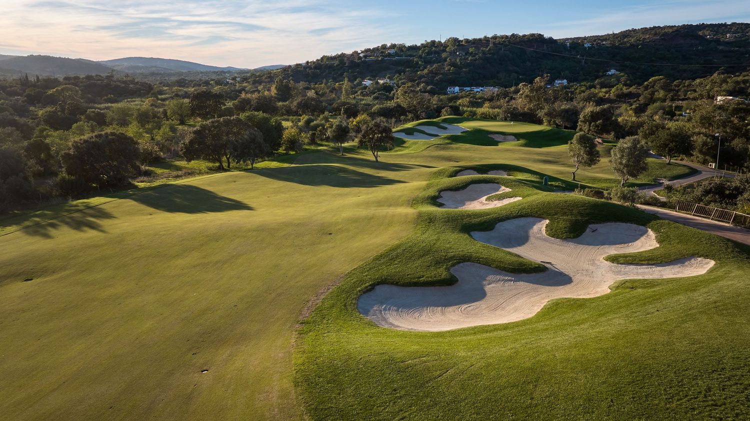Aerial view of a golf course featuring large sand bunkers, lush fairways, and surrounding hillside landscape.