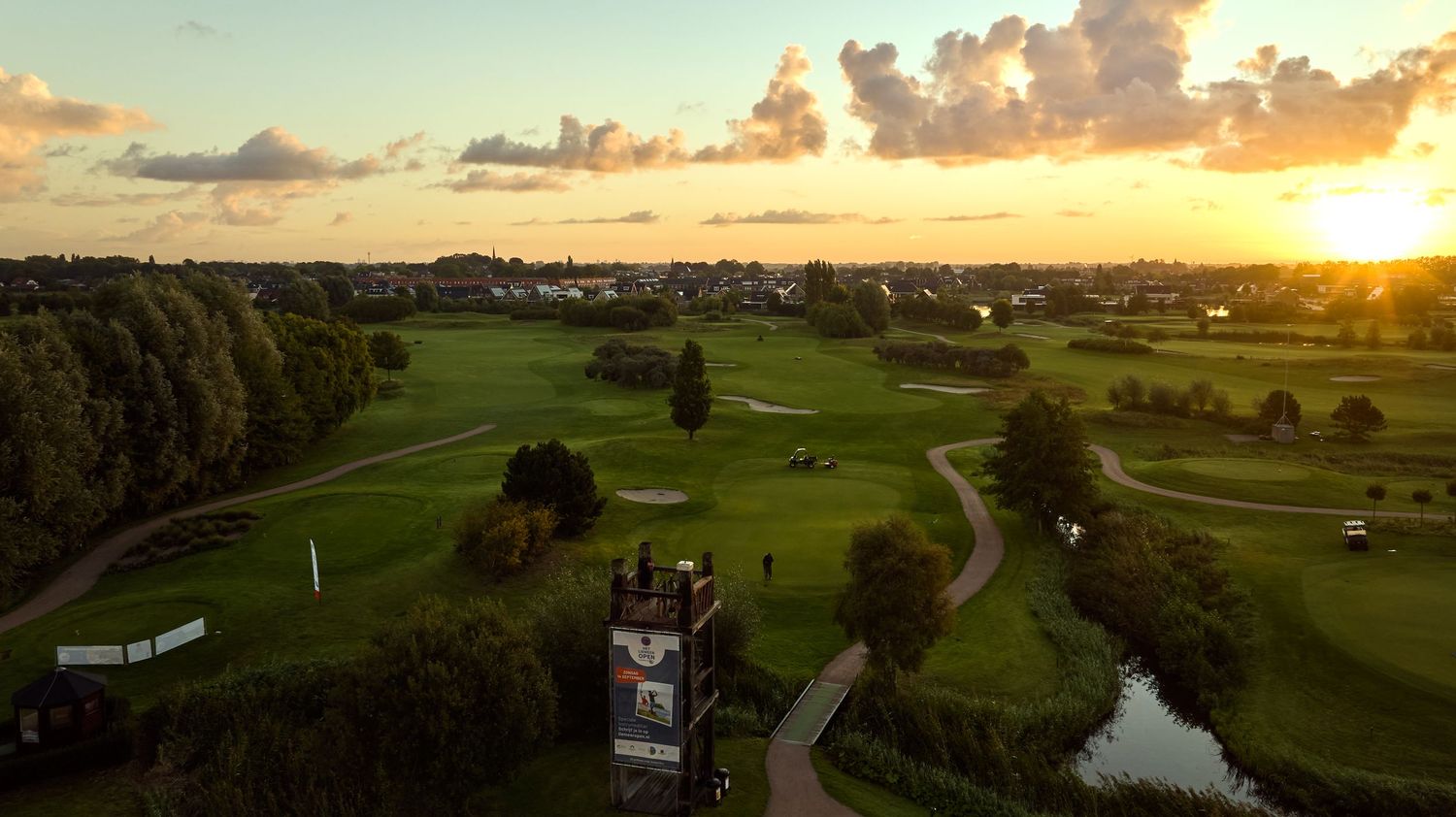 Aerial sunset view of a lush green golf course with a historic tower landmark surrounded by trees and urban landscape.