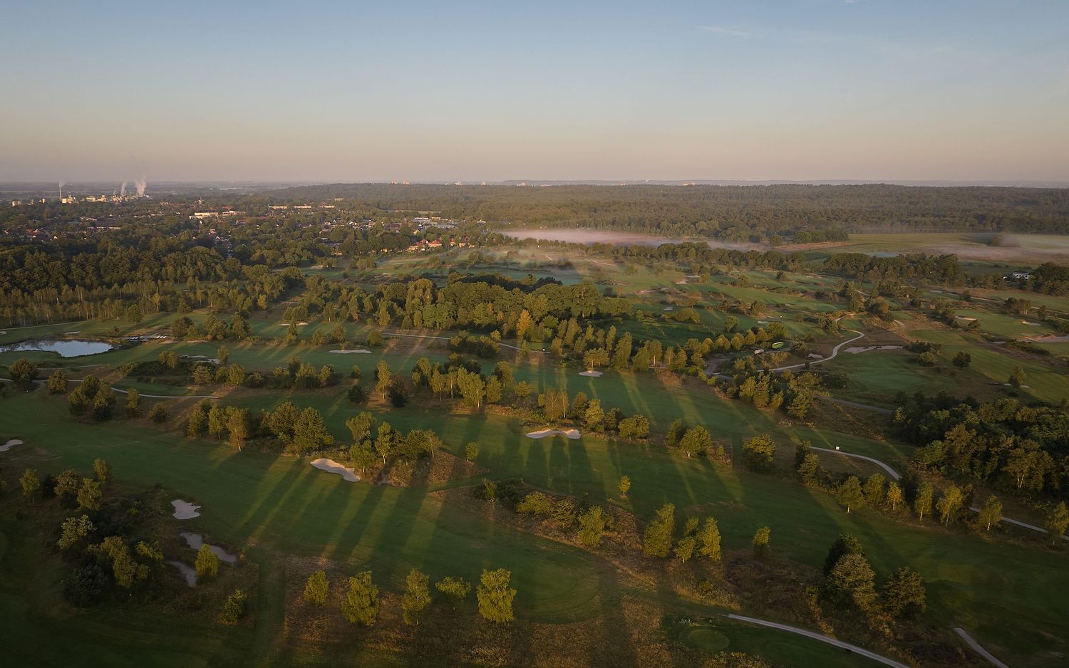 Wide aerial view of a vast golf course with tree-lined fairways, water features, and a city skyline in the distance.