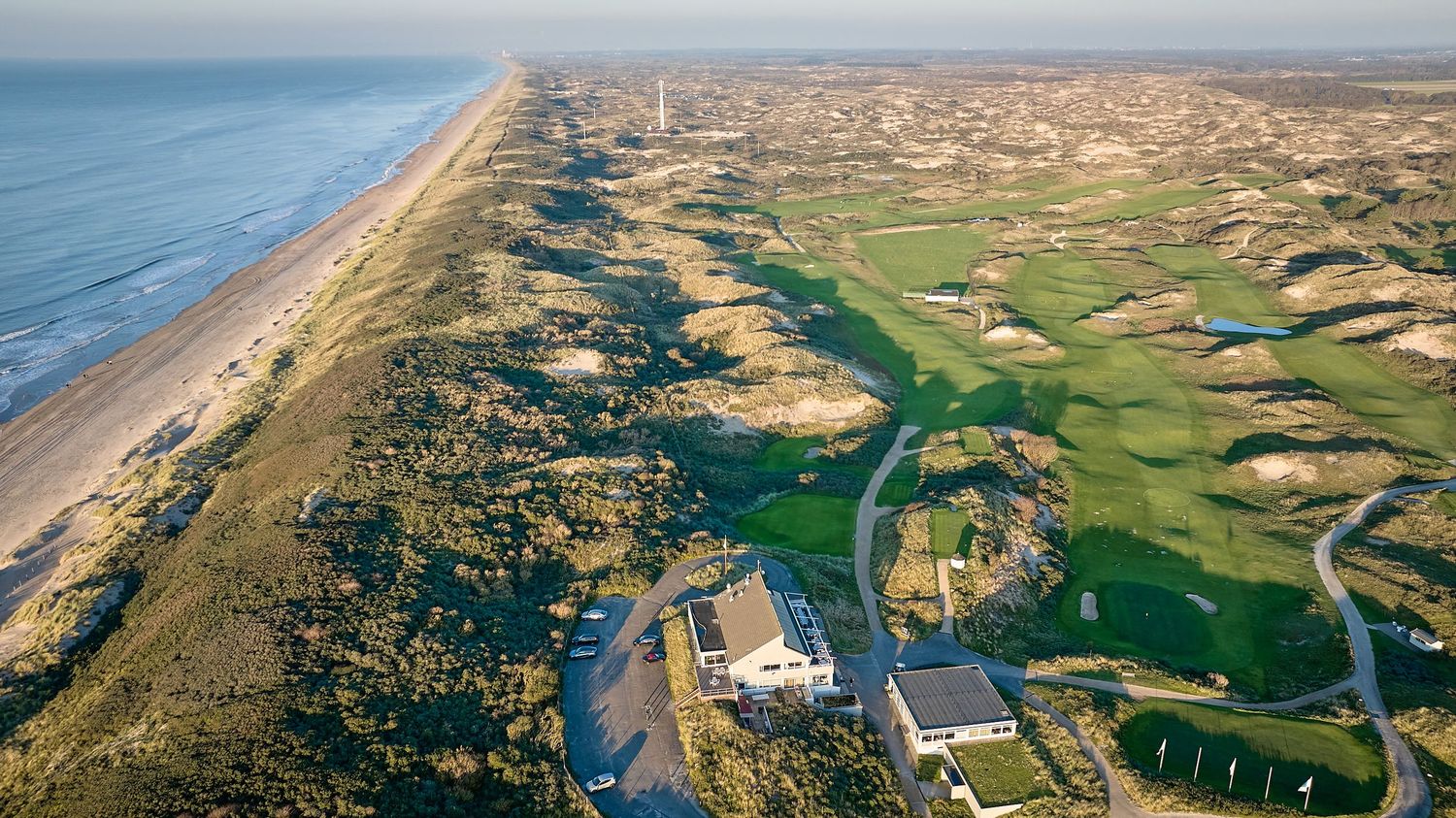 Aerial view of a seaside links golf course with sandy dunes, green fairways, a beach, and ocean coastline.