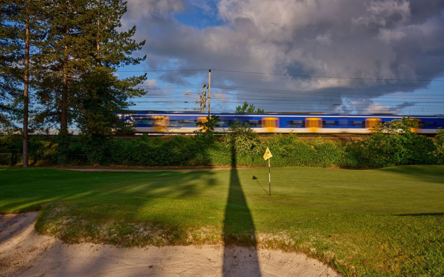A speeding blue train passes alongside a golf course green with bunker and flag in golden light.