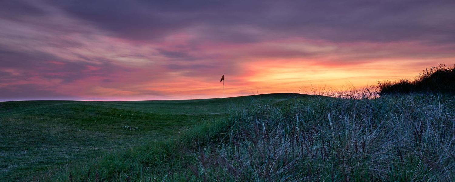 A golf course flag silhouetted against a vibrant purple and orange sunset sky over rolling greens.