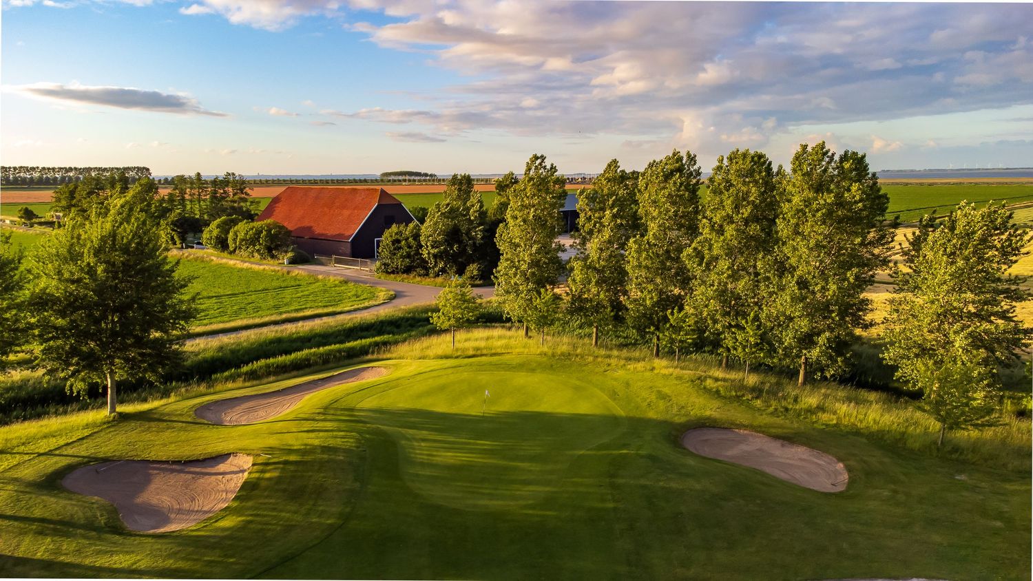 Golf course hole with sand bunkers near a red barn clubhouse under a vibrant blue sky.
