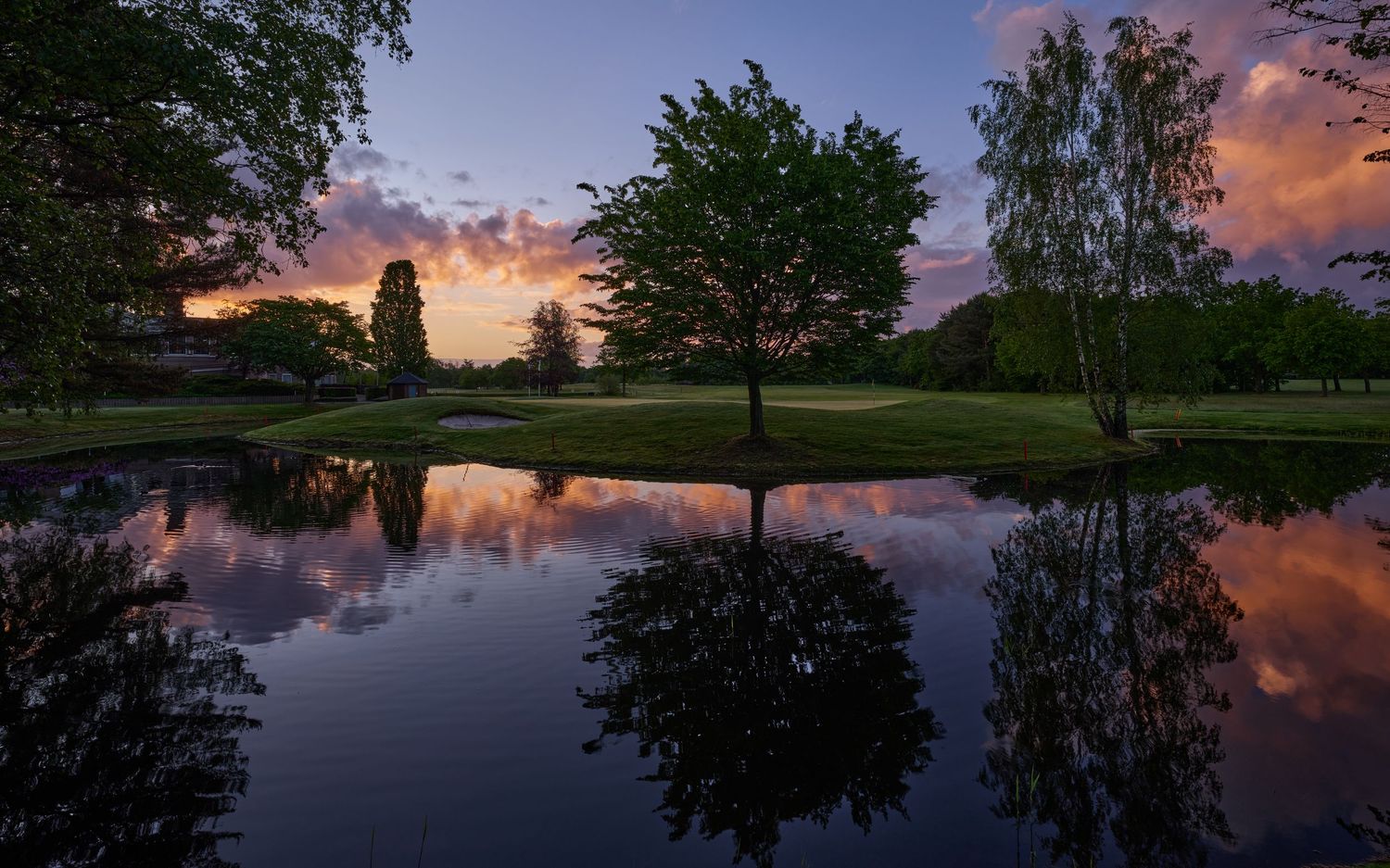 Tranquil golf course pond reflecting colorful sunset sky and lush green trees at twilight.