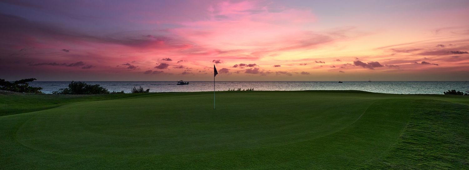 A golf green with a flag overlooks a calm ocean under a stunning pink and purple sunset.