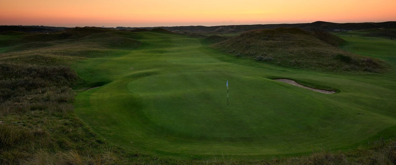 Scenic links golf course hole at dusk with lush green fairway, bunker, and dramatic sunset sky.