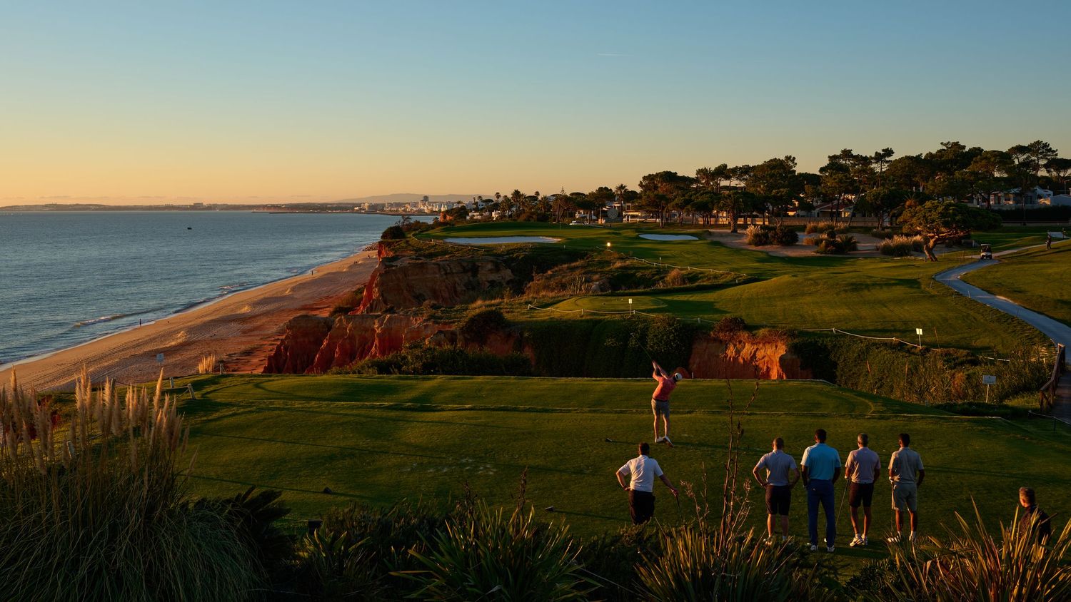 Golfers on a seaside course at golden hour with stunning coastal cliffs and ocean views in background.