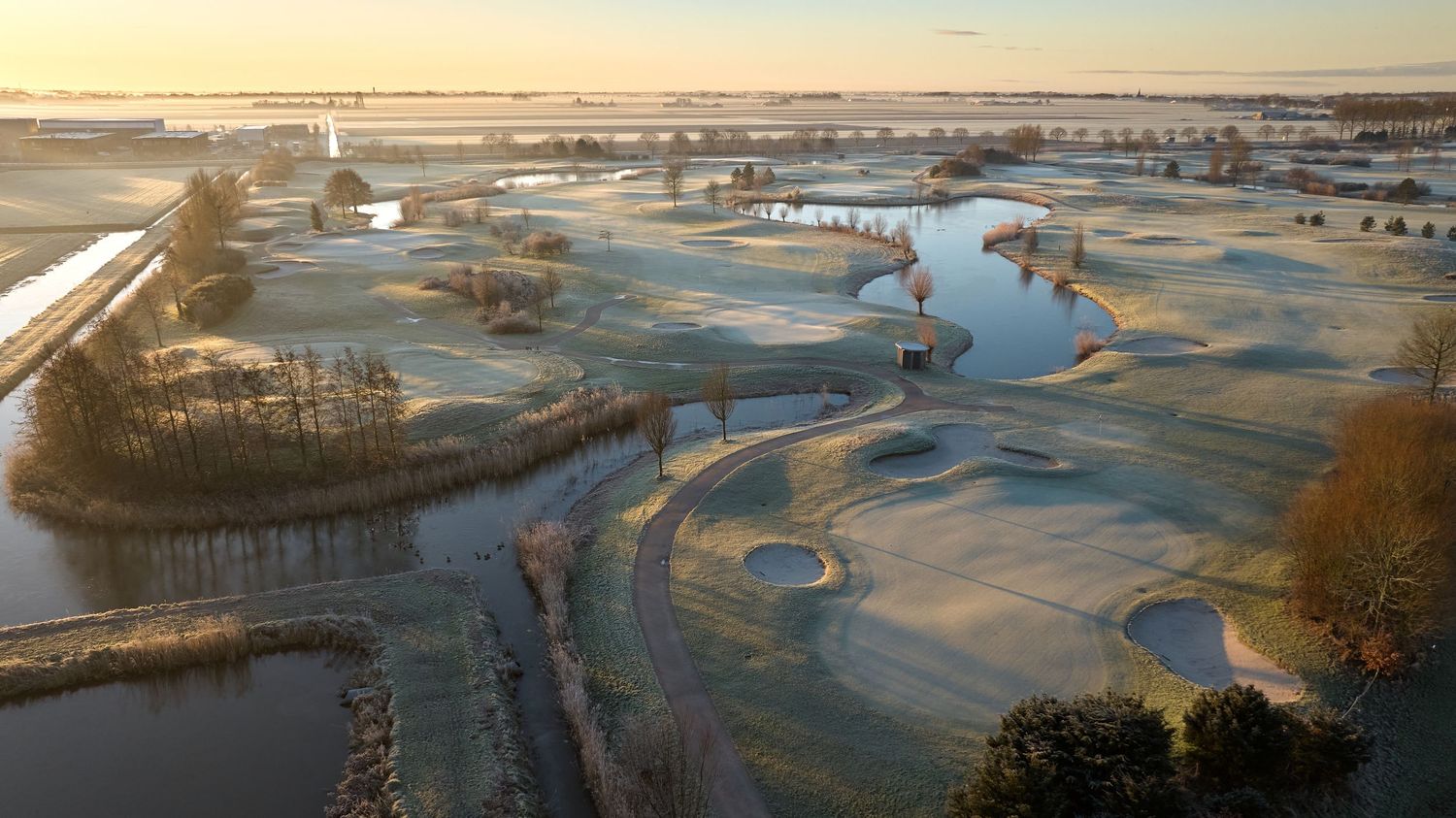 Aerial view of a frost-covered links golf course with water features and flat landscape at sunrise.