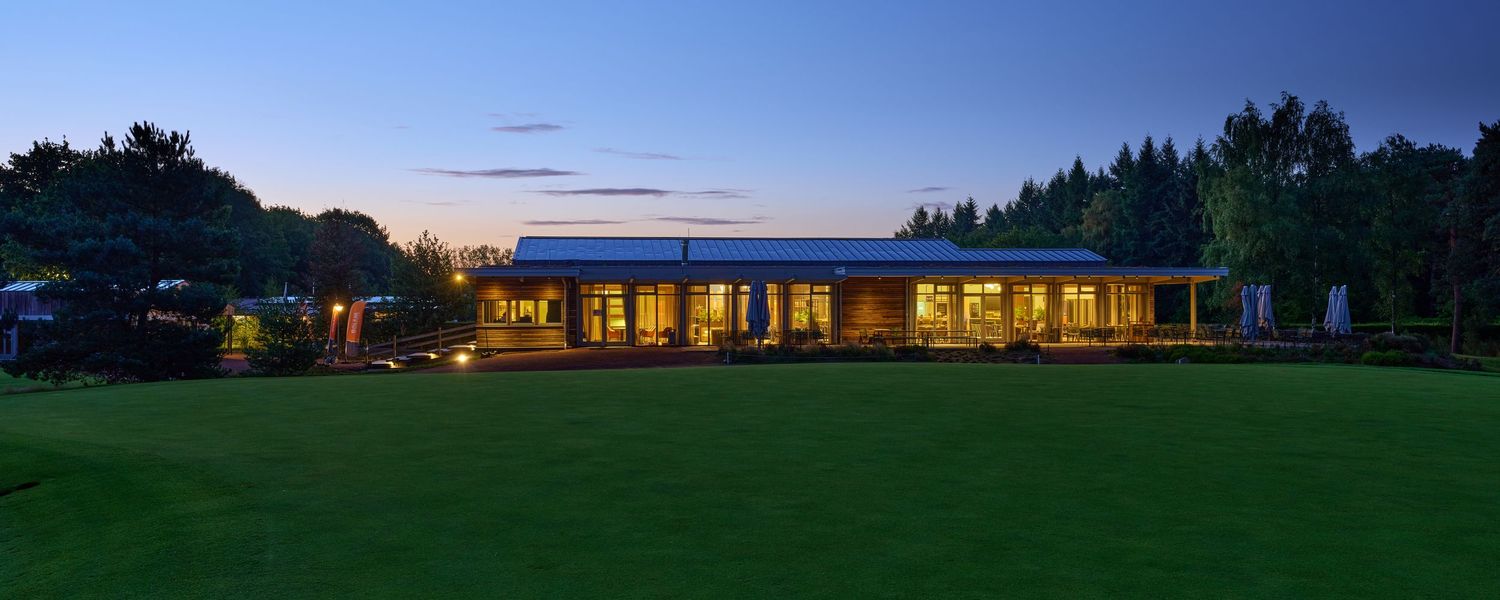 Elegant golf clubhouse illuminated at dusk surrounded by lush green lawn and tall trees at twilight.