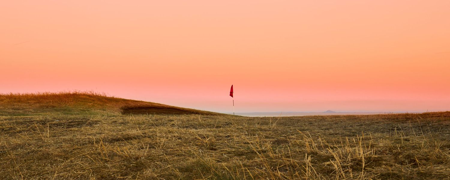 A lone golfer silhouetted against a stunning pink sunset sky on a links golf course with golden grass.