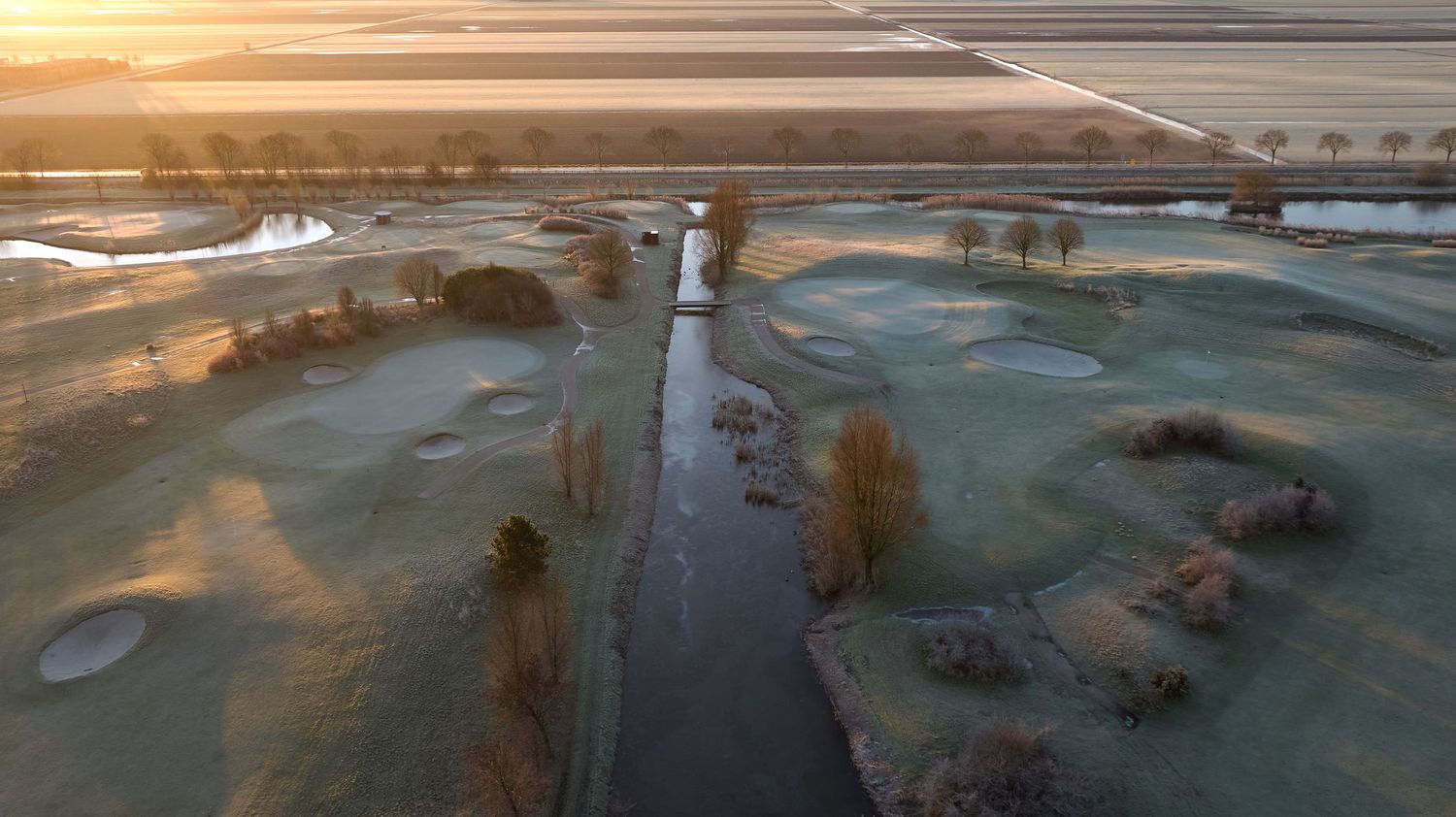 Aerial view of a frost-covered golf course with a canal and flat farmland at sunrise.