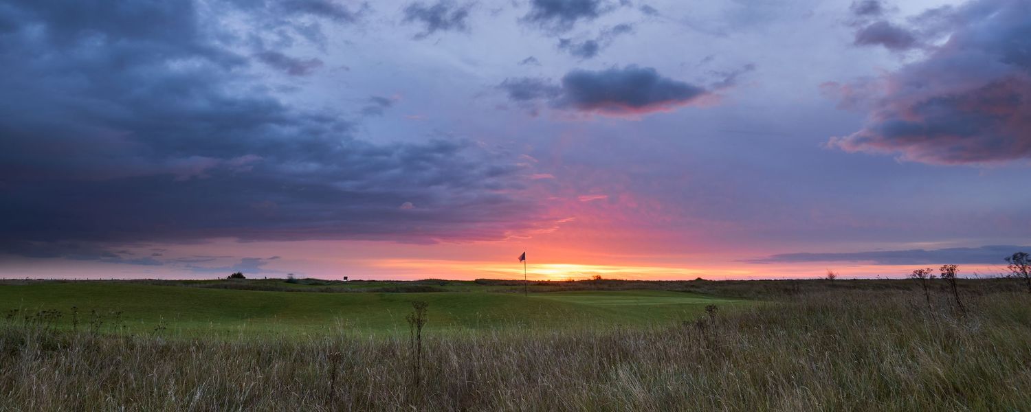 Wide panoramic view of a flat links golf course at sunset with glowing red sky and lone balloon.