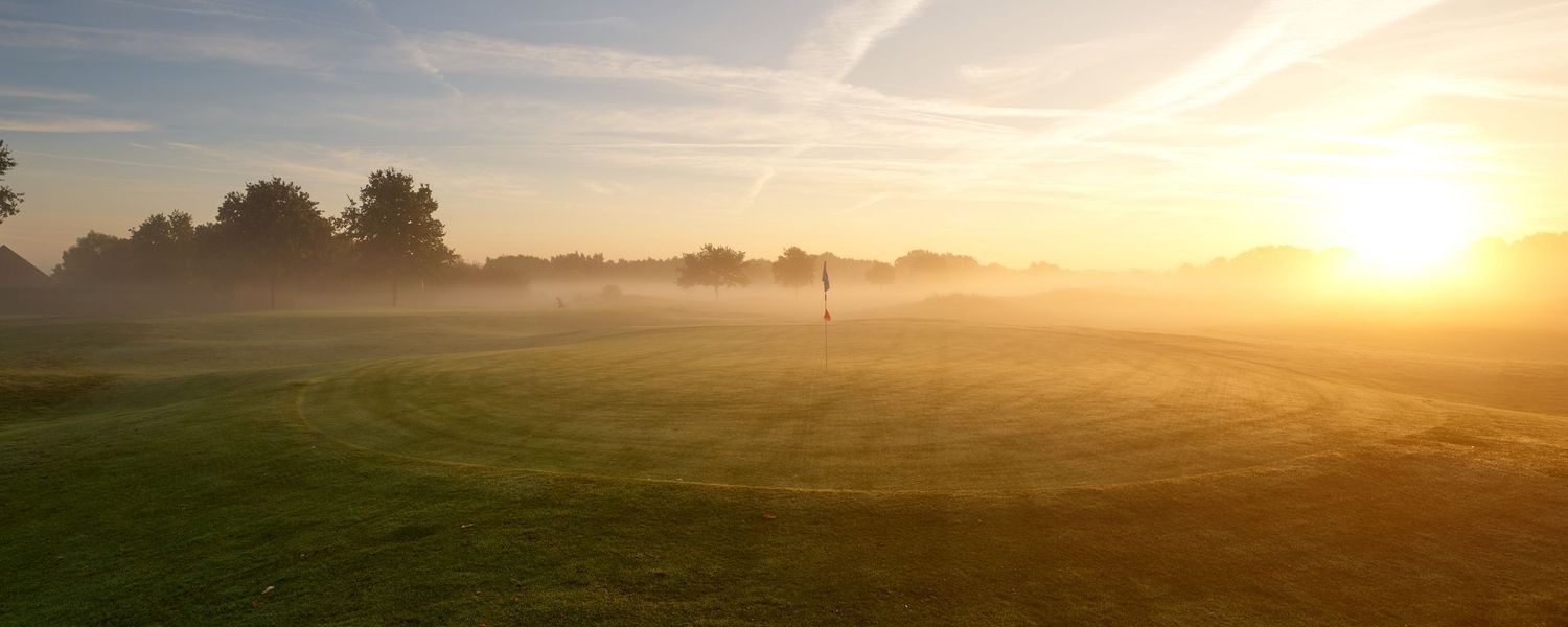 Misty sunrise over a golf course fairway with a lone flag pin glowing in golden morning light.