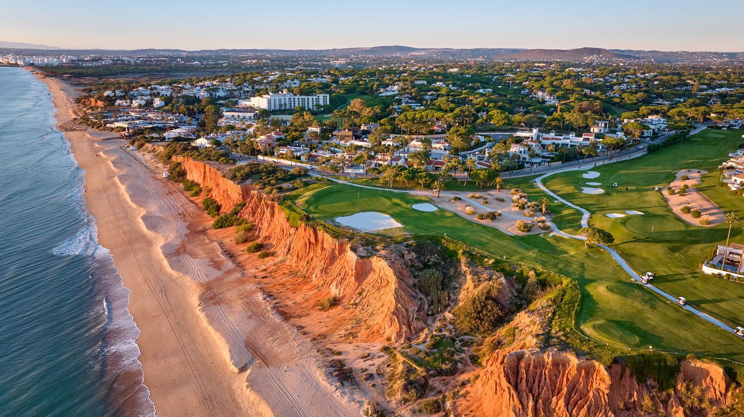Aerial view of a coastal golf course atop red cliffs beside a sandy Atlantic beach in the Algarve, Portugal.
