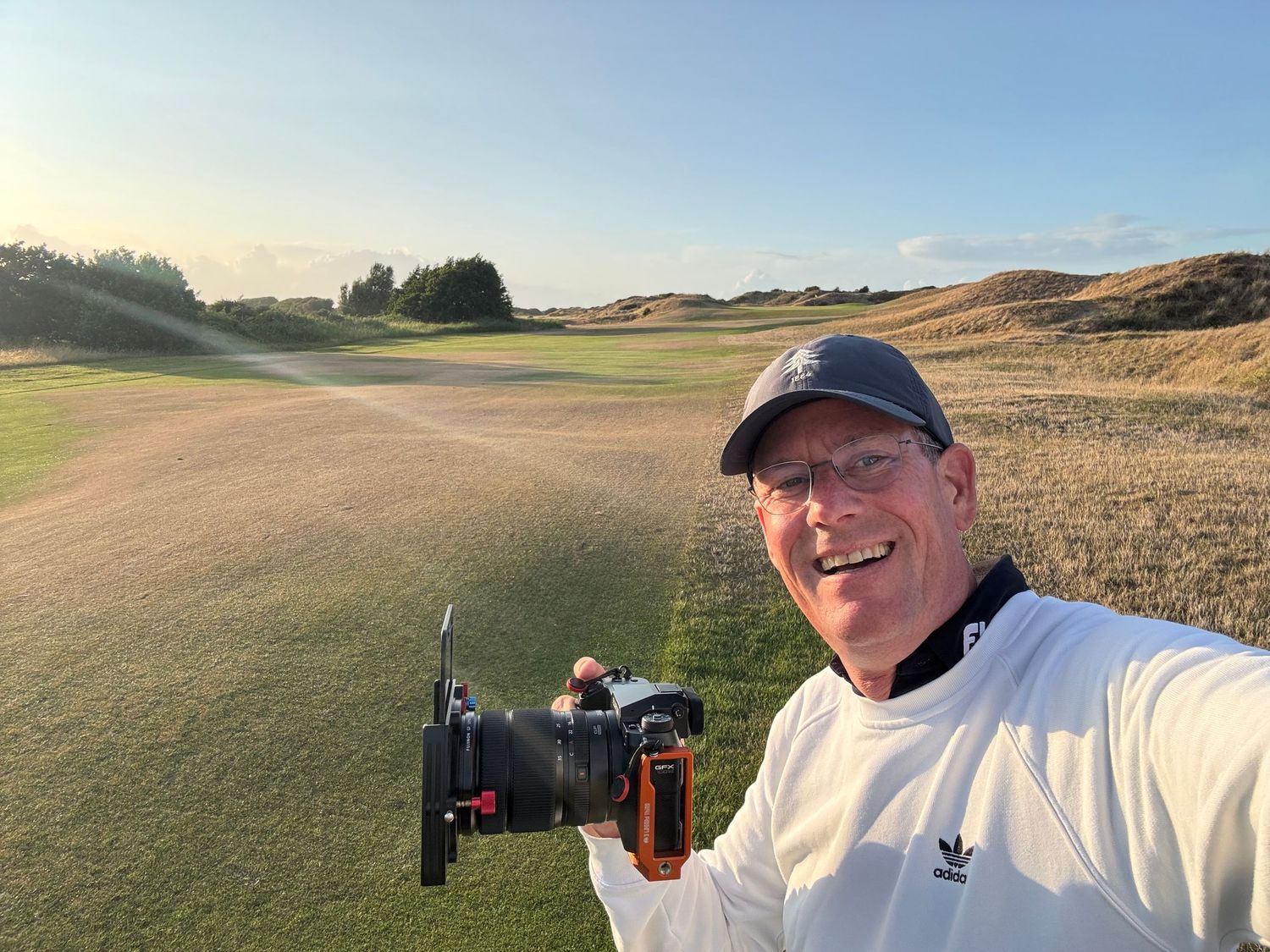 Smiling man taking a selfie with a camera on a scenic links golf course at golden hour.