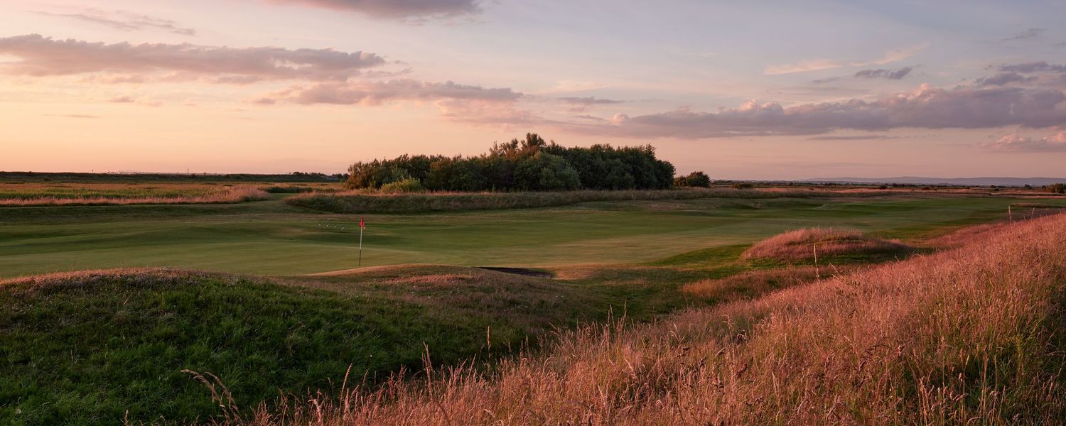 A links golf course stretches across open marshland under a dramatic pink and purple sunset sky.