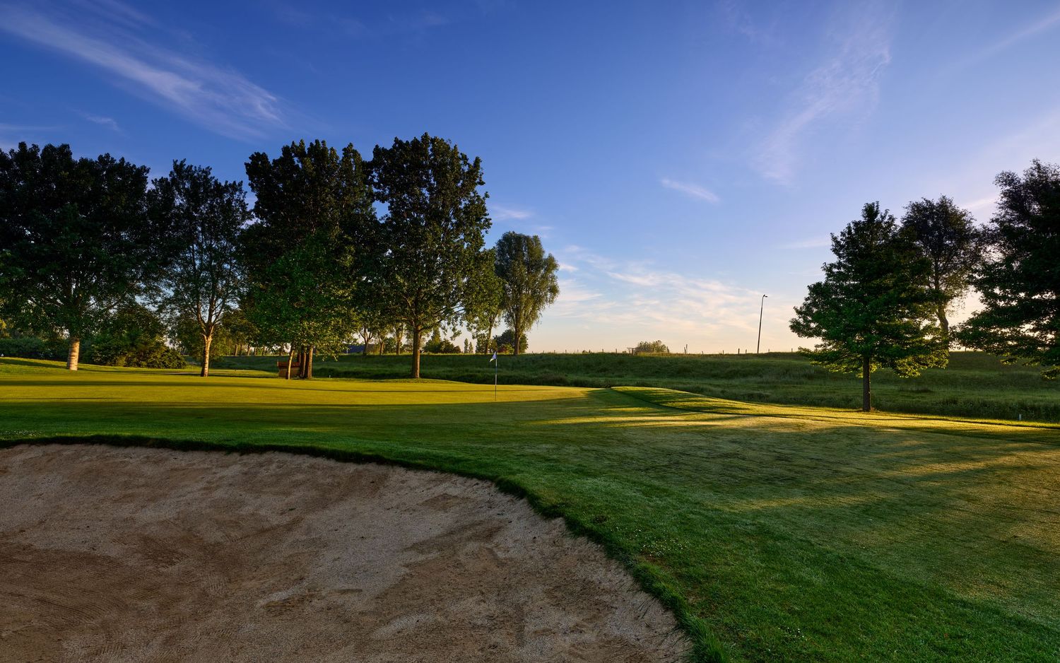 Sunny golf course fairway with sand bunker, lush green grass, and tall trees under a blue sky.