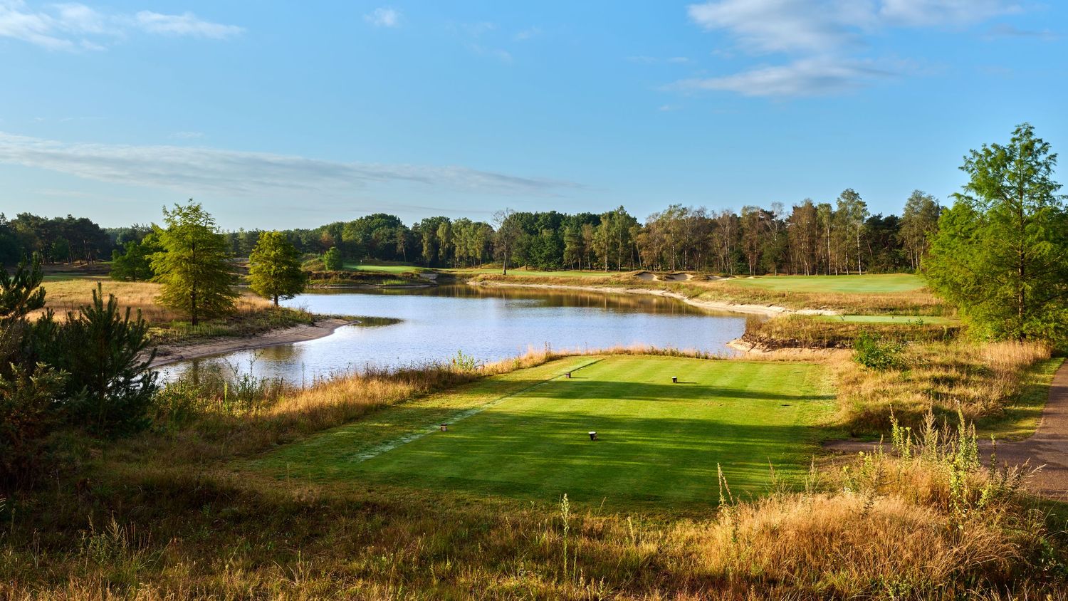 Scenic golf course with lush green fairways surrounding a tranquil pond amid autumn trees under a blue sky.