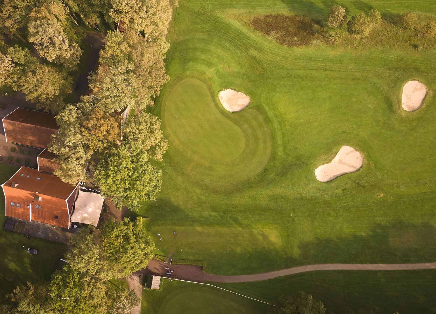 Aerial view of a golf course with sand bunkers beside a wooded area and red-roofed building.