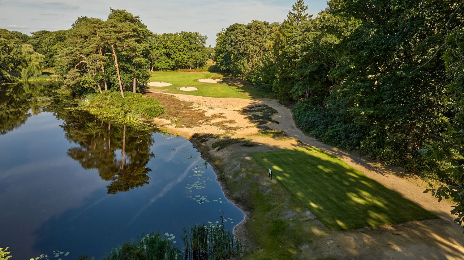 Scenic golf course hole with a reflective pond, sandy bunkers, lush green fairway, and tree-lined surroundings.
