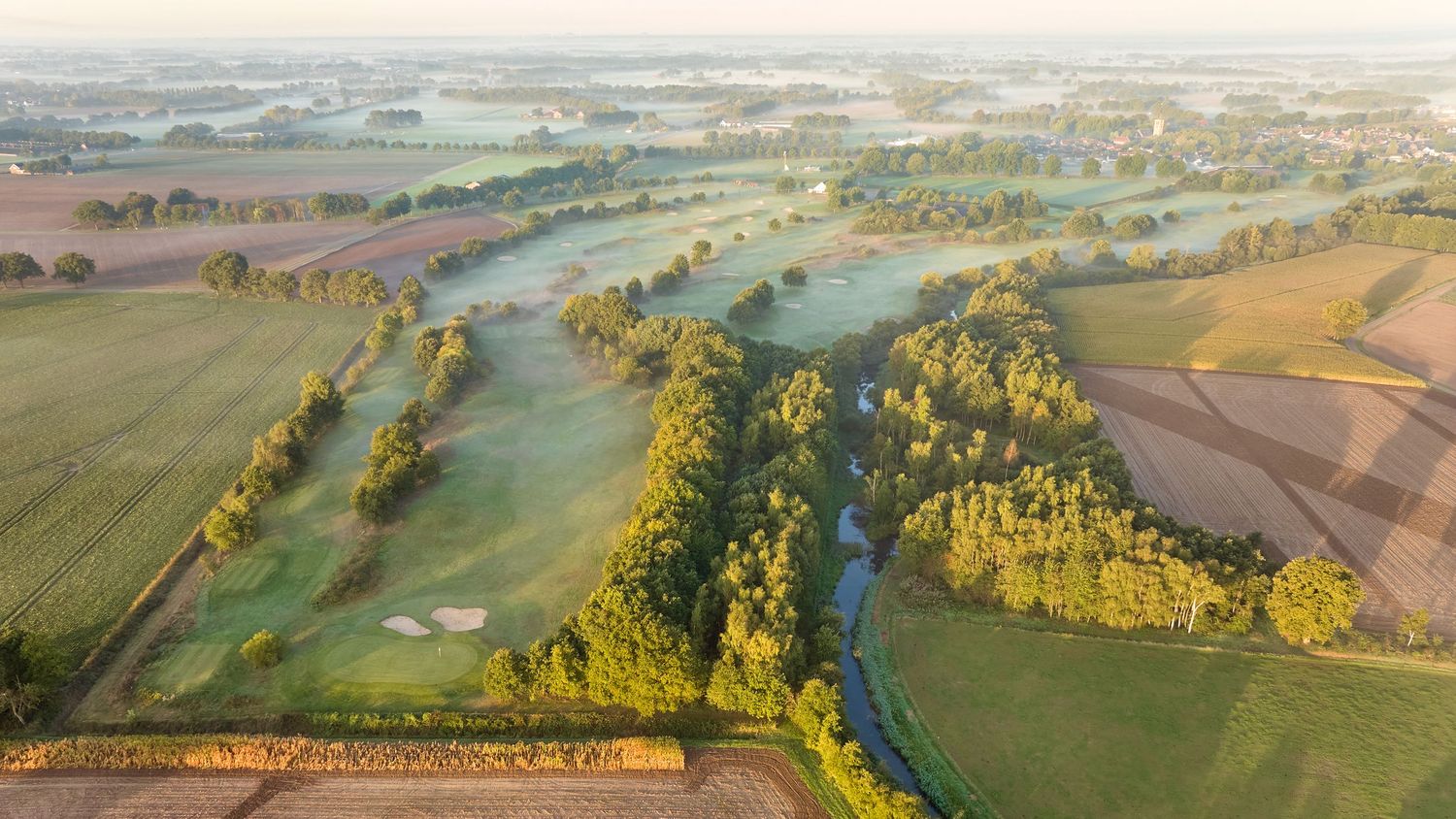 Aerial view of a scenic golf course winding through lush green fields and trees in morning mist.