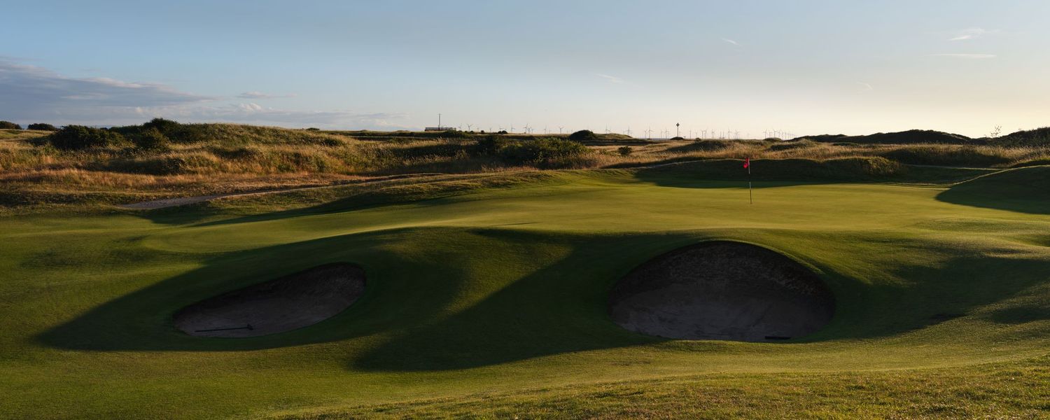 Links golf course with multiple sand bunkers, undulating green fairway, and blue sky with scattered clouds.
