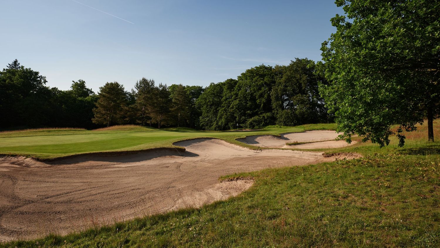 Large sand bunkers beside a golf course fairway surrounded by mature trees on a bright sunny day.