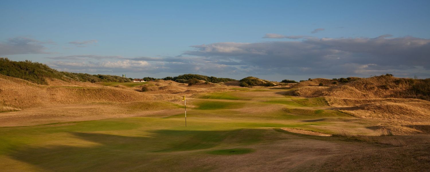 Links golf course with sandy dunes, green fairway, and yellow flag under blue sky.