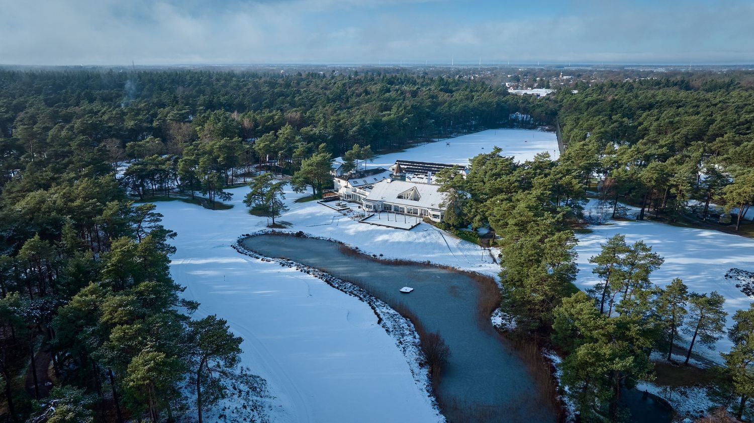 Aerial winter view of a snow-covered golf course surrounded by pine trees and a frozen lake.