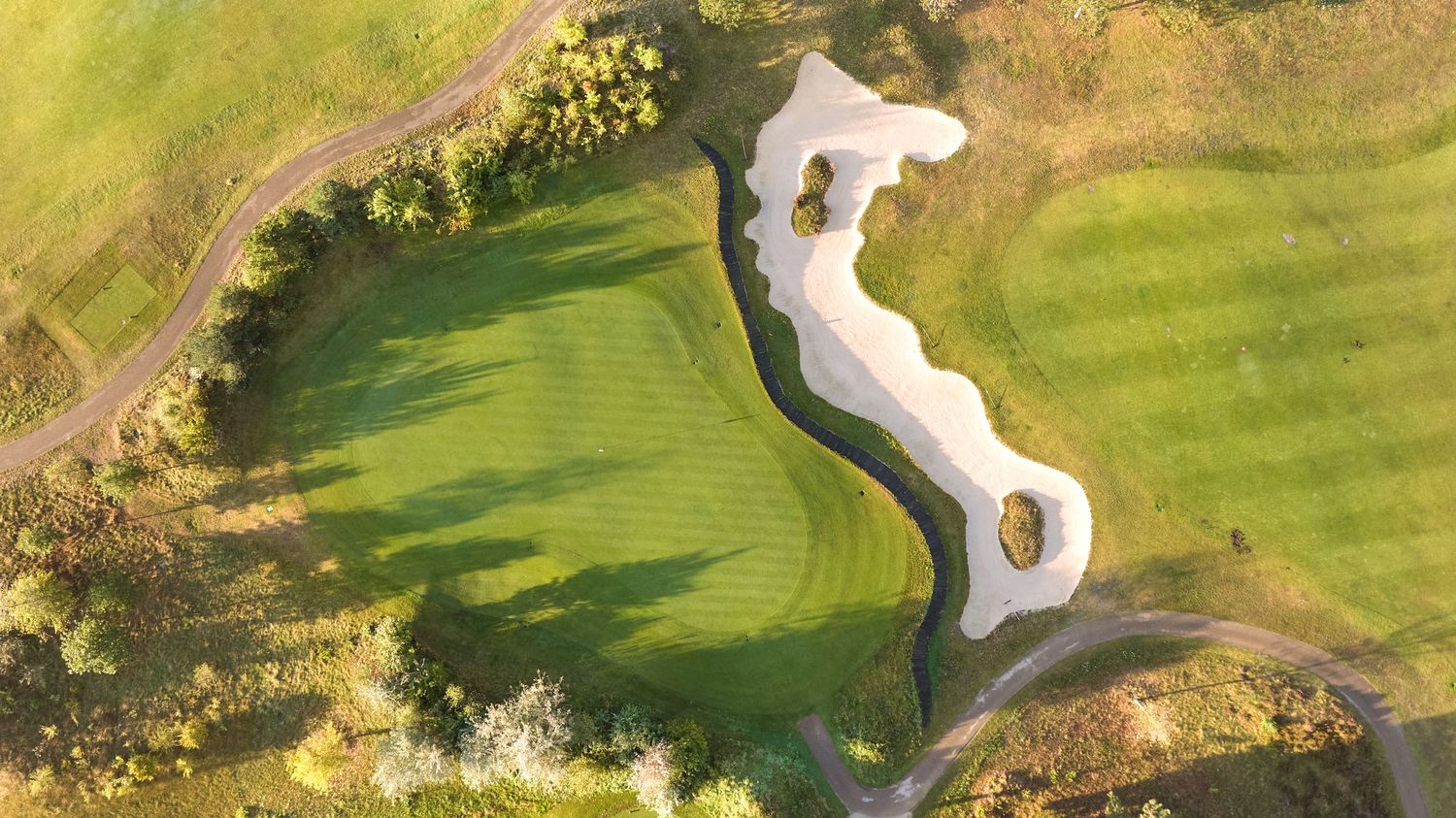 Aerial view of a winding golf course green with large sand bunkers and cart path at sunset.