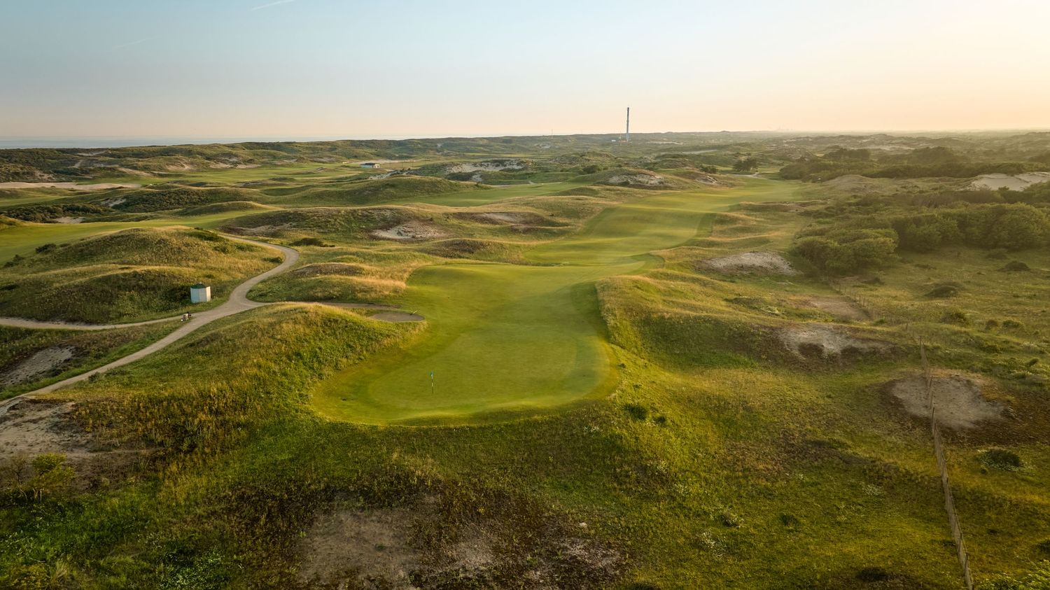Aerial view of a links golf course with undulating dunes, green fairways, and a distant wind turbine at sunset.