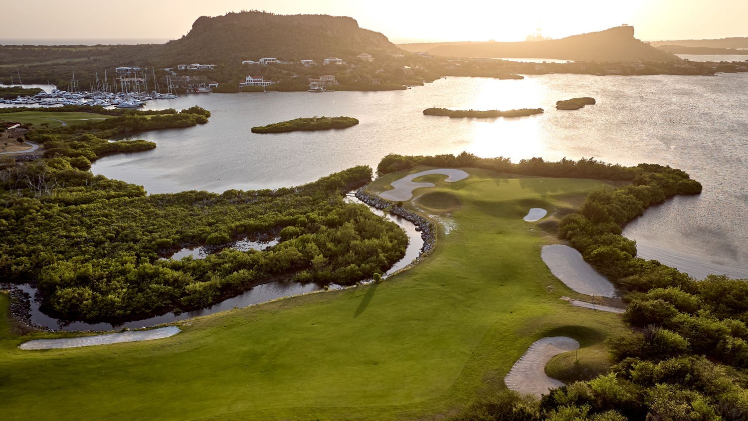 Aerial view of a golf course with sand bunkers winding through mangroves and lagoons at sunset.