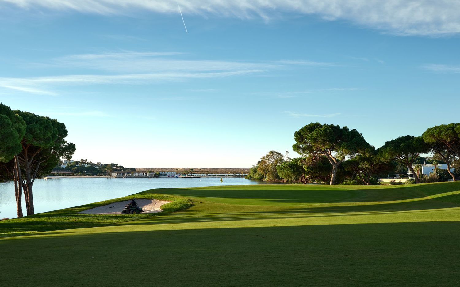 Scenic coastal golf course with lush green fairway, sand bunkers, and sparkling water views under clear blue sky.