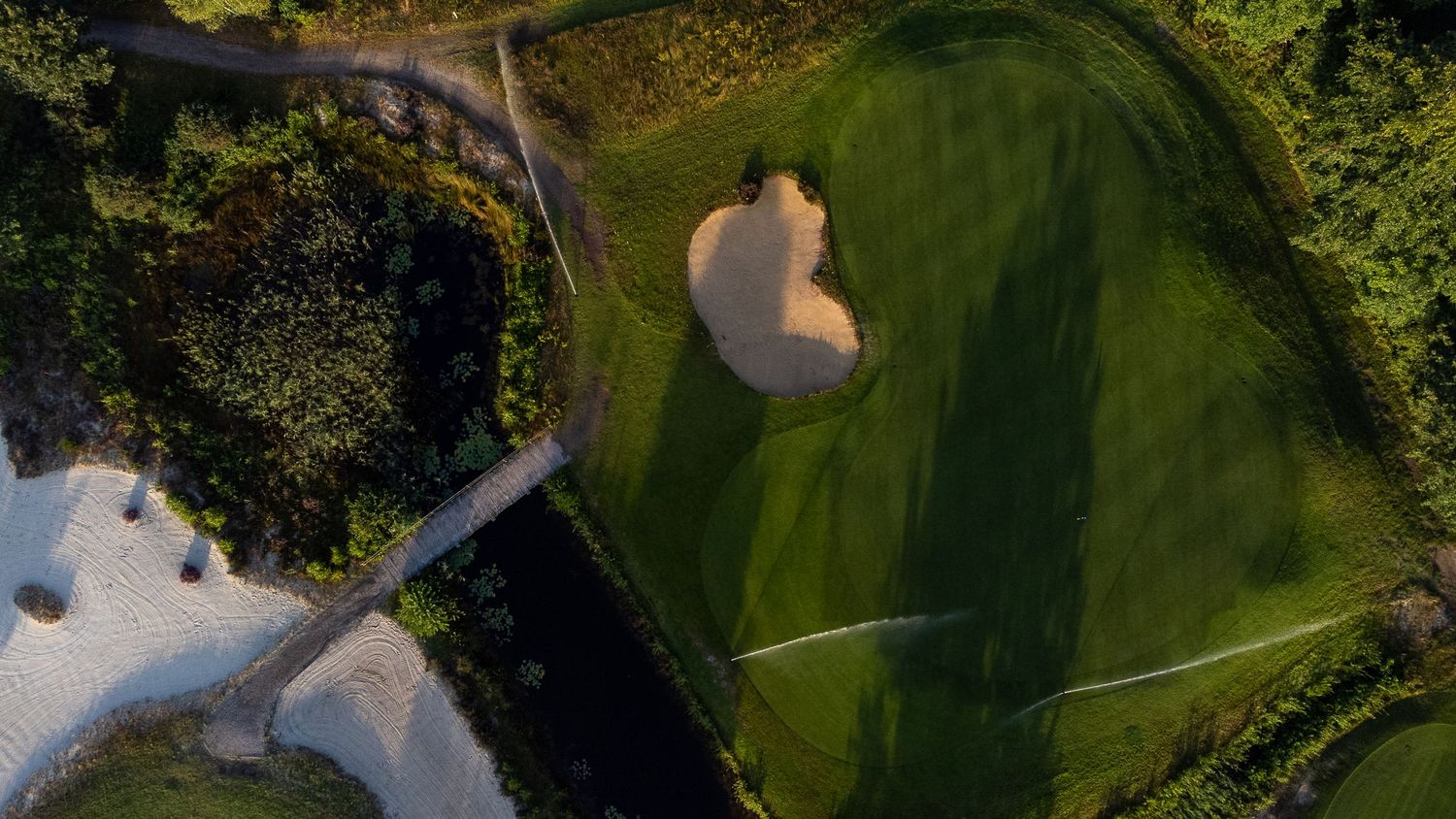 Top-down aerial view of a golf green with heart-shaped sand bunker beside a water hazard.
