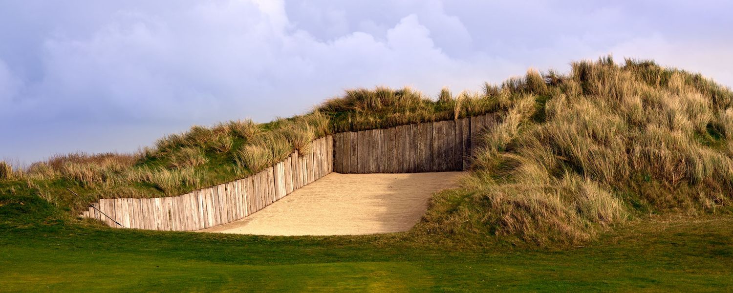 A wooden boardwalk path cuts through sandy dunes with tall grass on a links golf course.