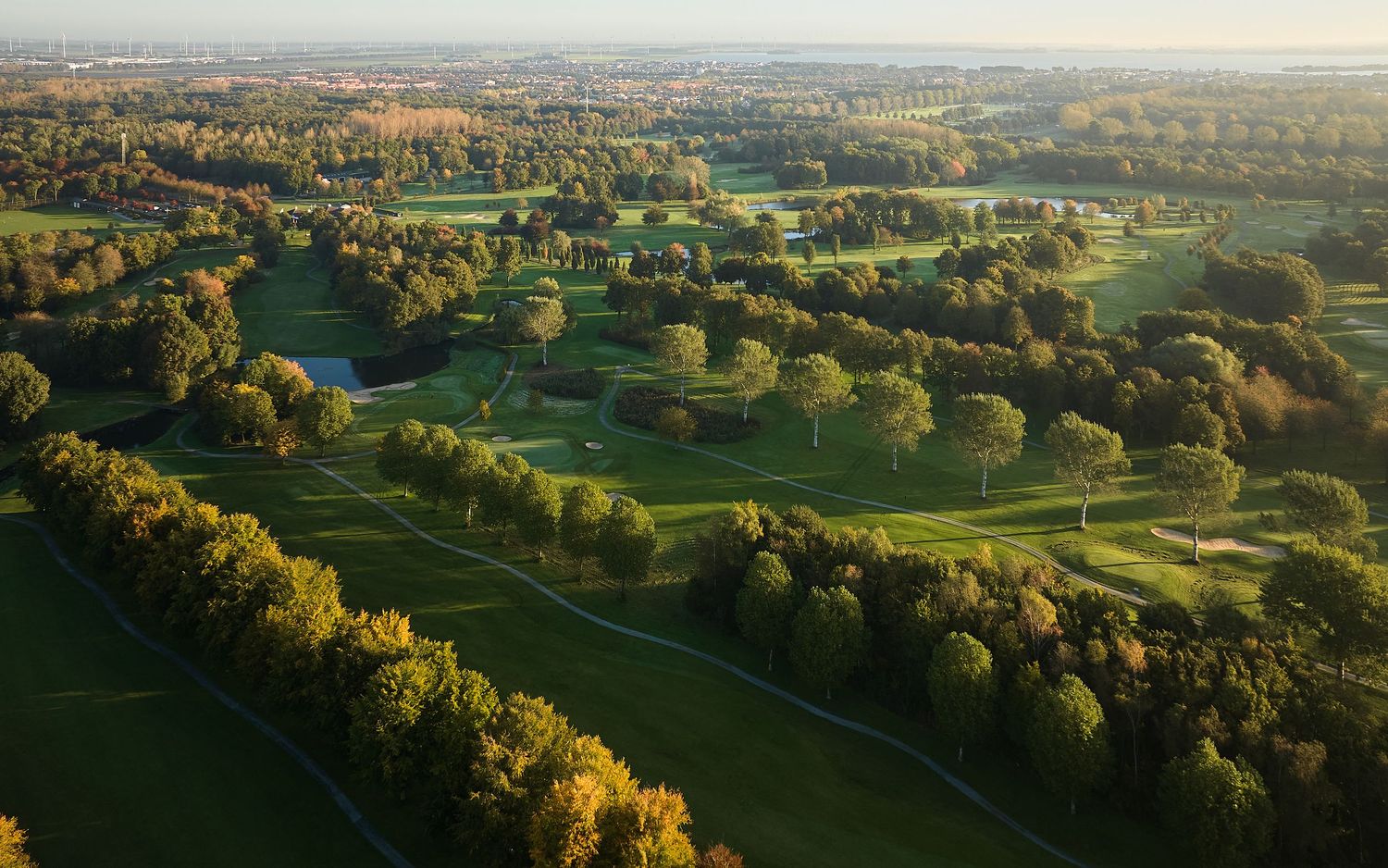 Aerial view of a lush golf course surrounded by autumn trees at golden hour.