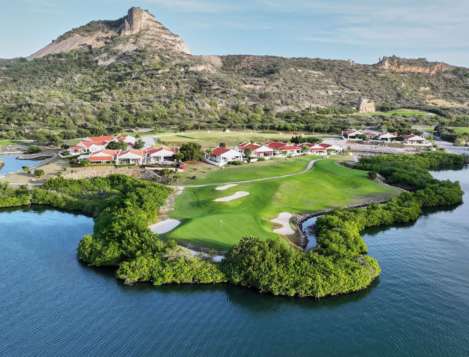 Aerial view of a tropical golf course surrounded by mangroves and water with a rocky mountain backdrop.