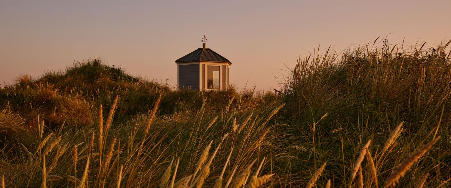 A small white chapel sits among golden grass dunes at sunset on a coastal landscape.