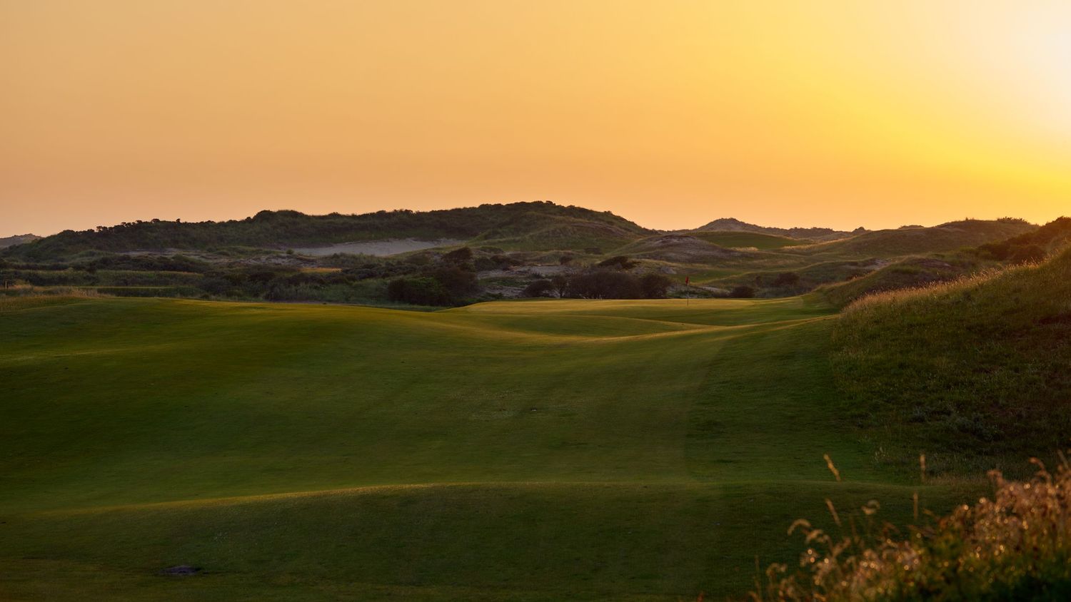Scenic links golf course at sunset with rolling dunes and vibrant orange sky on the horizon.