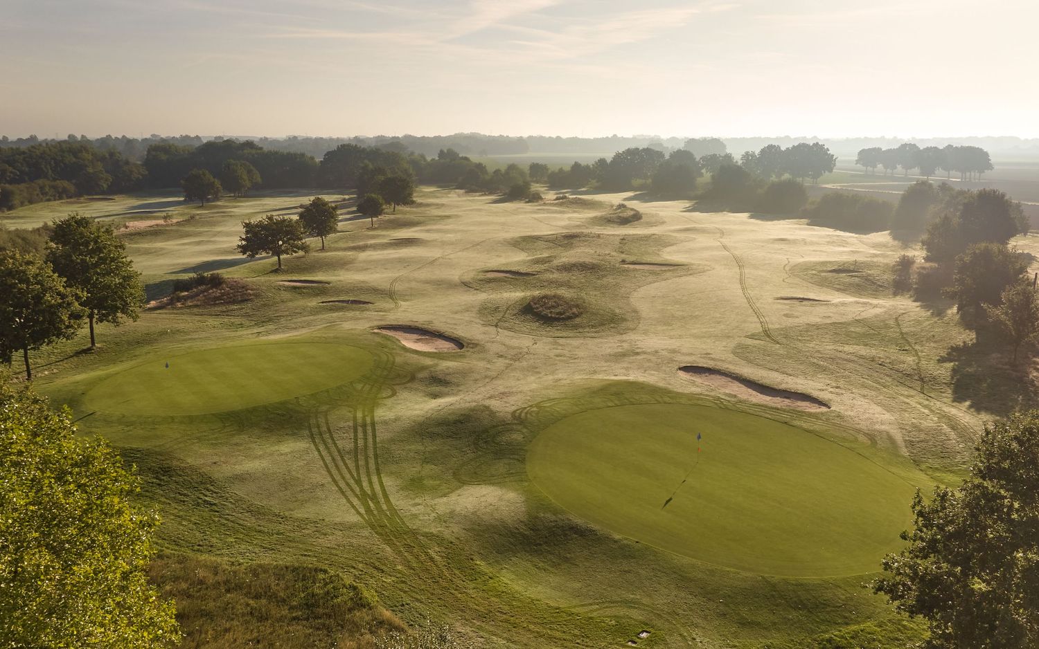 Aerial view of a golf course featuring undulating fairways, sand bunkers, and trees in morning haze.