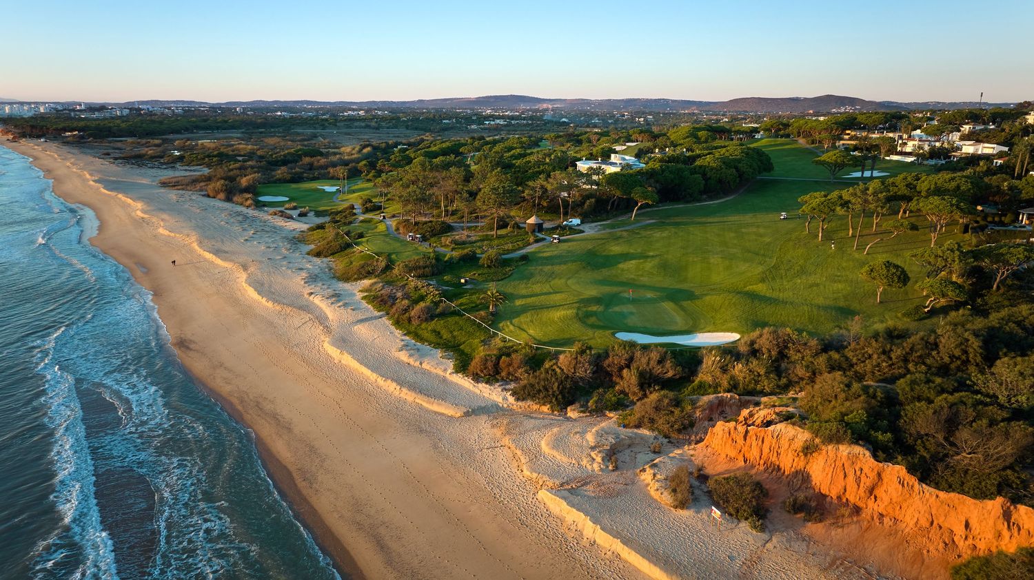 Aerial view of a golf course with red sandstone cliffs and golden sandy beach along the Algarve coastline.