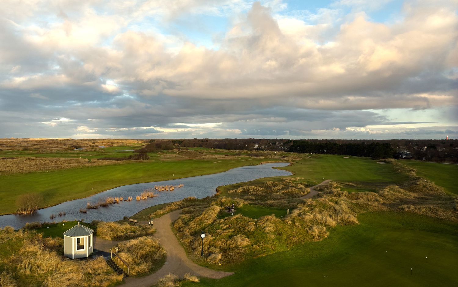 Aerial view of a links golf course with a small white building, winding river, and dramatic clouds.