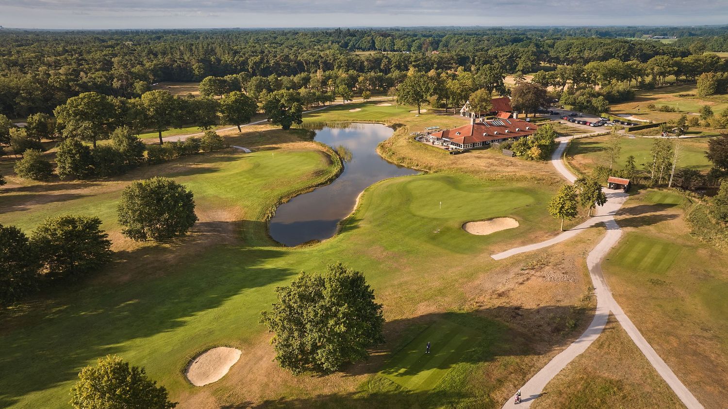 Aerial view of a golf course featuring a winding river, red clubhouse, manicured fairways, and surrounding woodland.