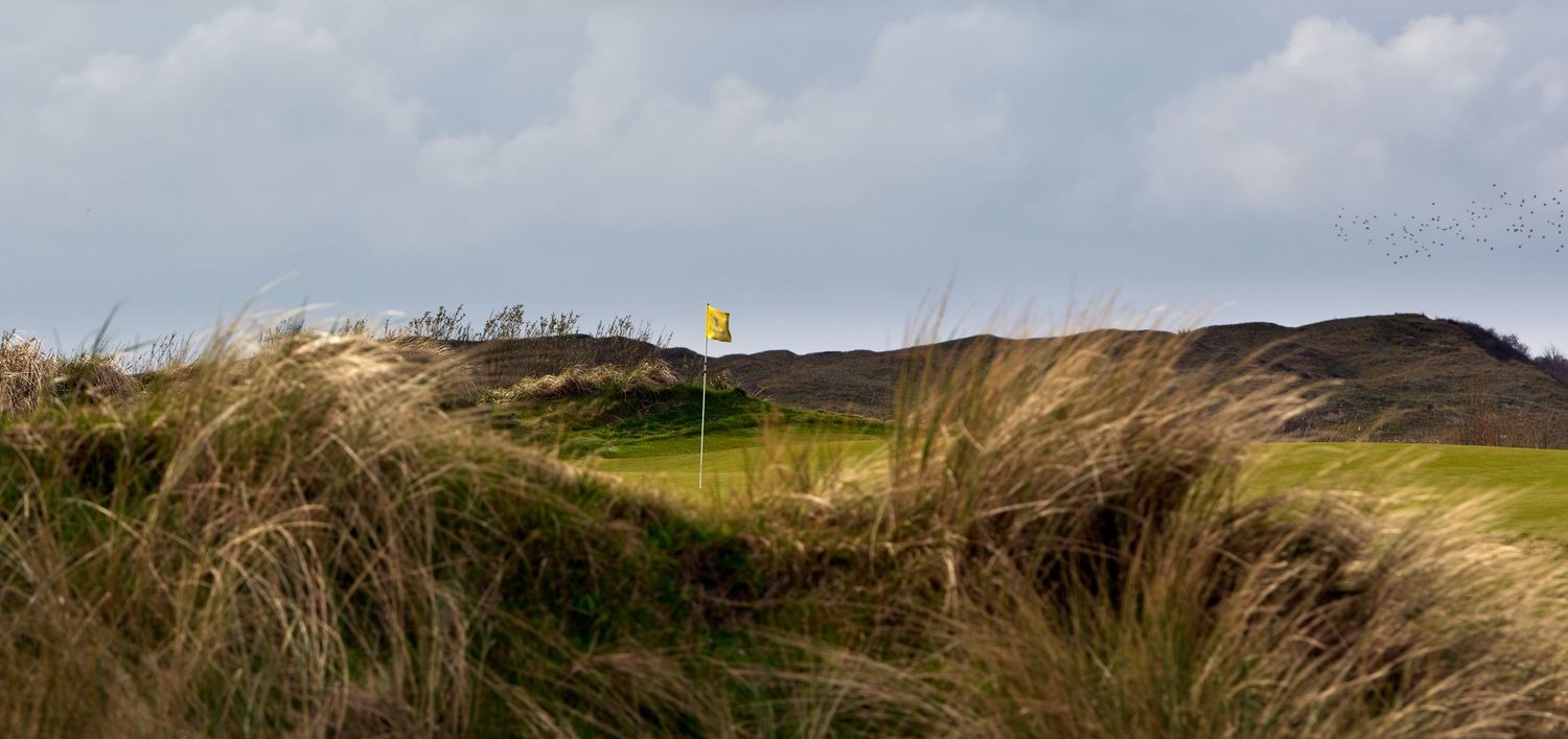 A yellow flag marks a golf hole nestled among windswept dunes and tall grass under a cloudy sky.